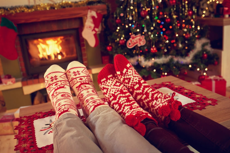 Two people wearing Christmas socks are warming themselves in front of the fireplace with a green Christmas tree next to them.