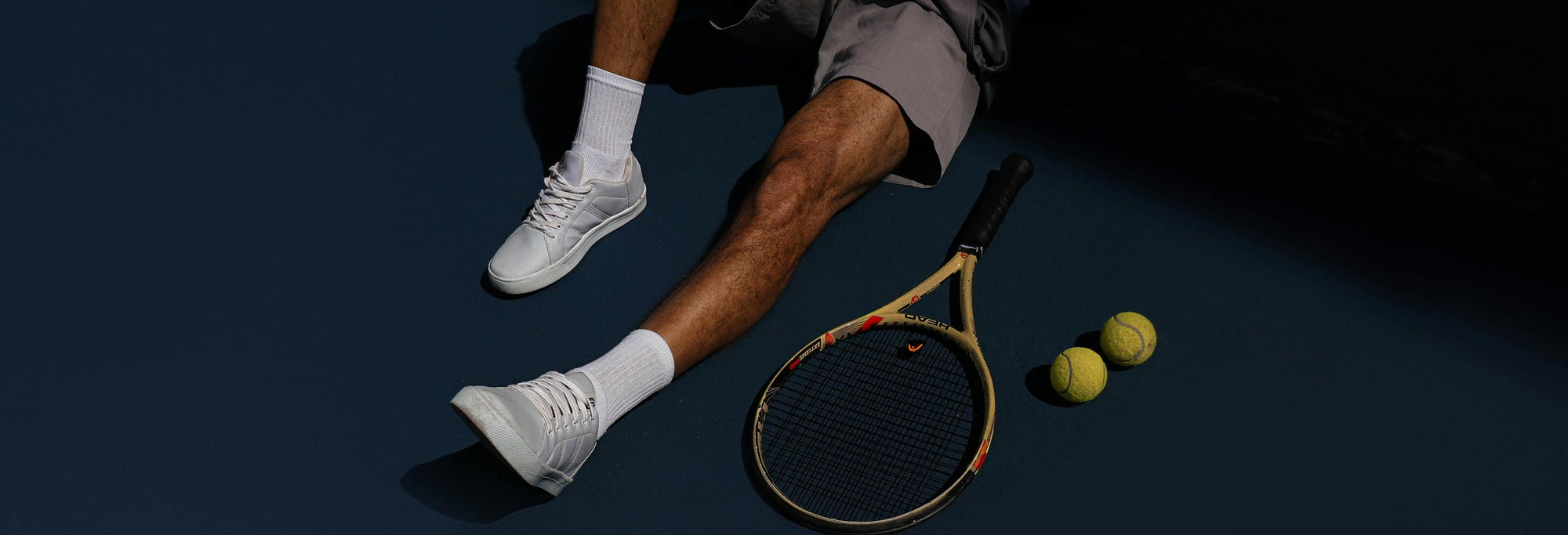 A person wearing white tennis socks and white sneakers sitting on a tennis court with a racket and tennis balls nearby.
